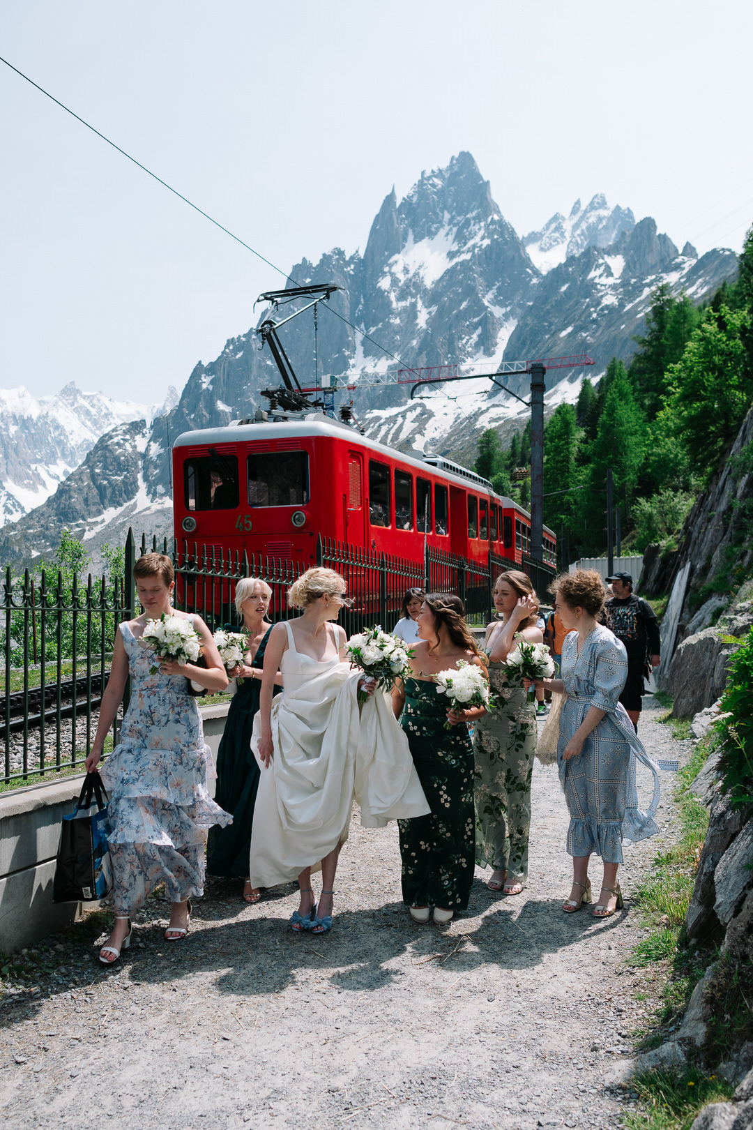 Bride and Bridesmaids walk along a mountain pass towards the wedding with a red mountain train in Chamonix behind them